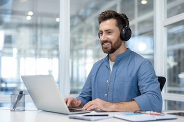 A smiling man wearing headphones works on a laptop in a modern office setting, looking happy.