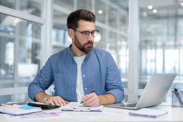 A man in a blue shirt sits at a desk, writing on paper. A laptop, calculator, and other office...