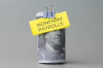 A rolled stack of US one-hundred dollar bills with a note "NONFARM PAYROLLS" attached. Economic indicator, labor market data, employment statistics concept.