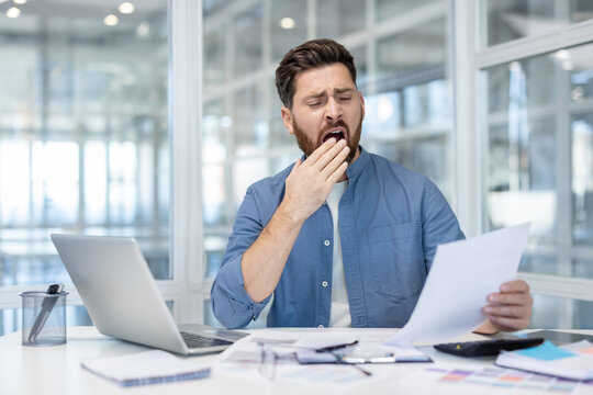A bored businessman yawns while reviewing documents at his desk, showing signs of fatigue and exhaustion. - Powered by Adobe
