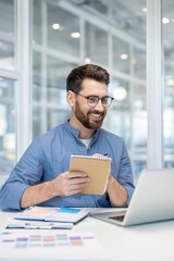 A smiling man with glasses takes notes while looking at his laptop at a modern office.