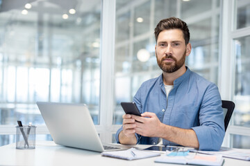 A confident businessman in a blue shirt uses his smartphone in a modern office setting, looking...