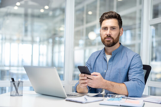 A confident businessman in a blue shirt uses his smartphone in a modern office setting, looking directly at the camera with a focused expression.