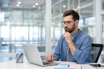 A pensive man in glasses works on a laptop in a modern office setting, pondering a problem or task.