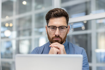 A thoughtful man with a beard wearing glasses looks intently at a laptop screen, hands clasped, in...