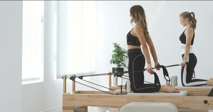 Two beautiful young women in black sportswear doing pilates reformer exercises for hands with double loop straps in modern bright studio.                                                       