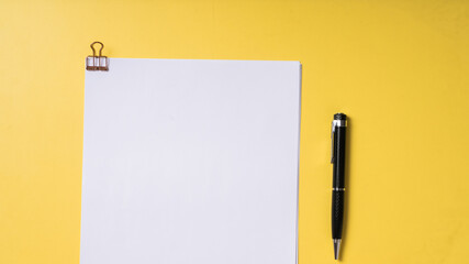 A black pen and a stack of white papers clipped with a rose gold binder clip on a solid yellow background, captured in a clean, top-down flat lay style