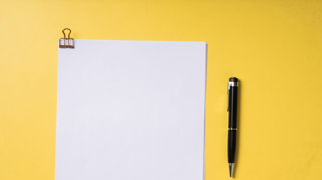 A black pen and a stack of white papers clipped with a rose gold binder clip on a solid yellow background, captured in a clean, top-down flat lay style