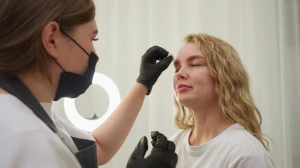 Beautician wearing black gloves gently wipes eyebrow area of blond client with cotton pad in well-lit beauty salon setting, preparing skin for treatment or makeup application