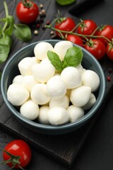 Tasty mozzarella cheese balls in bowl, basil, peppercorns and tomatoes on black table, above view