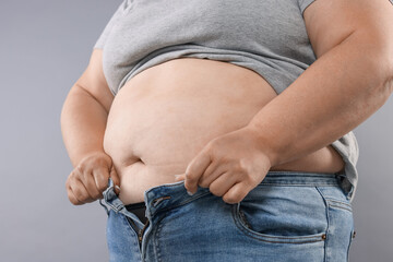 Overweight woman trying to put on tight jeans against grey background, closeup
