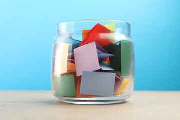 Colorful paper pieces in glass jar on wooden table against light blue background
