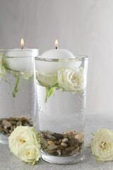 Burning candles in glasses with water, stones and rose flowers on light grey table, closeup