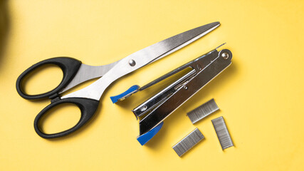 A metal stapler, black-handled scissors, and four staple pieces are arranged on a bright yellow background, creating a vibrant composition of essential office tools