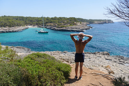 young athletic man looking from the shore at a sailing yacht at anchor in a beautiful sea bay, summer vacation