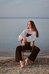 Young woman in casual attire sits on chair at serene beach, embodying calmness and introspection....