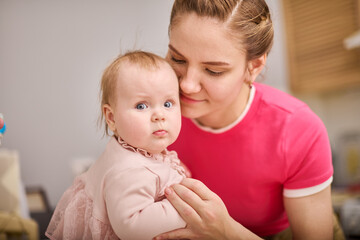 Young mother with brown hair holds baby girl in pink dress indoors. Warm lighting creates intimate, cozy environment. Baby curious expression contrasts with mothers gentle smile
