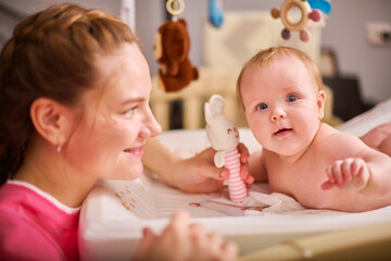Smiling adult mother with brown hair plays with joyful baby girl on changing table. Soft lighting...