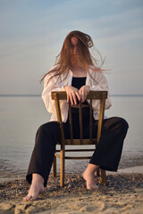 Young woman sits on chair facing calm sea, symbolizing solitude and introspection. Her relaxed posture and flowing hair evoke serene, contemplative mood at dusk