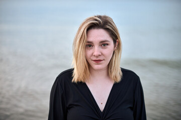Young woman with blonde hair and serene expression stands by sea. Overcast sky and gentle waves...
