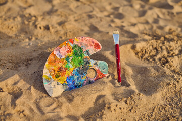 Vibrant artist palette and paintbrush lie on sandy beach, blending creativity with nature. Setting...