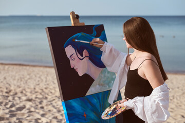Young female artist in black dress paints portrait on canvas set against serene seaside. Scene...