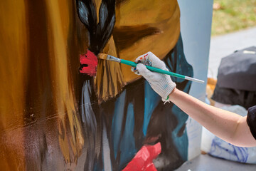 Focused view of adults hand painting mural outdoors. Gloved hand holds brush applying vivid red and...