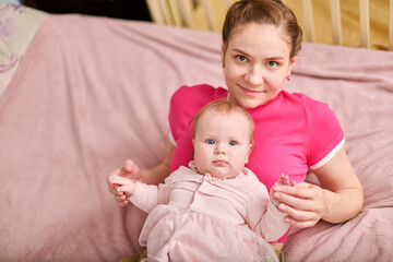 Young adult female with brown hair holding adorable baby girl in pink outfit. European ethnicity, both smiling indoors on soft pink blanket with wooden crib nearby