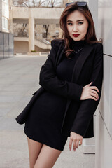 A confident woman in a black dress and blazer poses outdoors against a modern wall