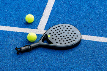 A black padel racket and two neon yellow balls lie on a blue artificial turf court with intersecting white lines, illustrating a sporty and energetic match setup