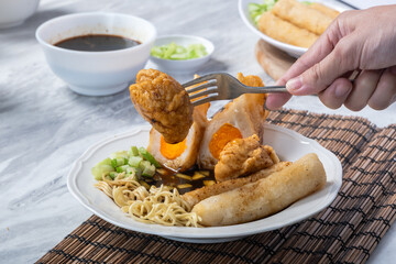 Asian hand picking fried pempek with fork, served with noodles, cucumber, and cuko sauce on a white plate atop a bamboo mat background, showing traditional Indonesian delicacy
