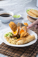 Asian hand picking fried pempek with fork, served with noodles, cucumber, and cuko sauce on a white plate atop a bamboo mat background, showing traditional Indonesian delicacy