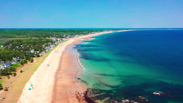 Drone captures vibrant beaches in Saco, Maine. Tourists walk, play, and relax on sunny sand as Atlantic waves roll in. Vacation homes line the coastline, showcasing New England&rsquo;s summer seashore charm