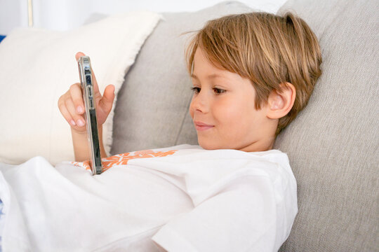 Smiling child boy wearing a white t-shirt lying on a couch using smartphone. Technology concept. - Powered by Adobe