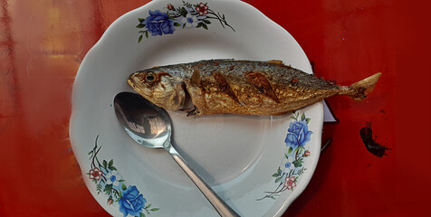 A single fried fish served on a floral-patterned plate with a metal spoon. Simple traditional meal presentation on a red table background