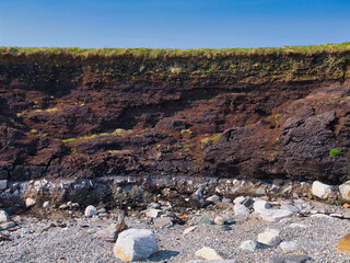 Layers of dark peat above a rocky beach topped with green grass in Heoga Ness, Yell, Shetland, UK.
