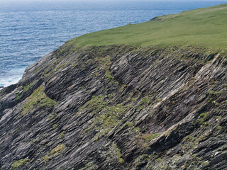 Rugged coastal cliffs at St Ninian's Isle, Shetland meet the ocean, showing rock strata and a green landscape under a clear sky. Coastal cliffs of the Colla Firth Formation - Semipelite and psammite
