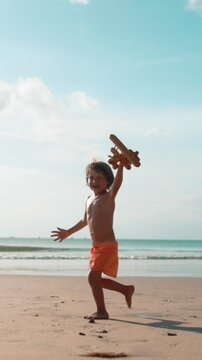 Happy little boy running along seaside with wooden toy airplane in hand, laughing and playing as if plane is flying. Slow motion scene of childhood joy, freedom, imagination and summer adventure on
