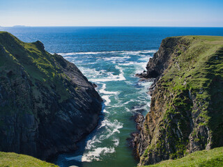 Stunning view of a rugged inlet cutting through cliffs on Unst's west coast, Shetland, bathed in vibrant sunlight.
