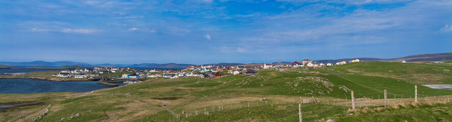 A panoramic view of the coastal community of Hamnavoe in Shetland, UK, nestled between rolling green hills and a serene bay under a clear blue sky.
