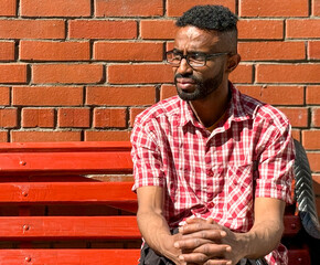 Portrait of a young man of African-American nationality wearing glasses with a beard sitting on a bench. A black guy in a plaid shirt on the background of a brick wall. Portrait of a black man. 