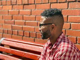 Portrait of a young man of African-American nationality wearing glasses with a beard sitting on a bench. A black guy in a plaid shirt on the background of a brick wall. Portrait of a black man. 