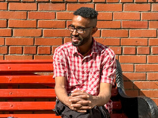Portrait of a young man of African-American nationality wearing glasses with a beard sitting on a bench. A black guy in a plaid shirt on the background of a brick wall. Portrait of a black man. 