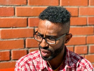 Portrait of a young man of African-American nationality wearing glasses with a beard sitting on a bench. A black guy in a plaid shirt on the background of a brick wall. Portrait of a black man. 
