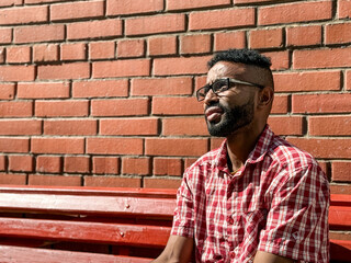 Portrait of a young man of African-American nationality wearing glasses with a beard sitting on a bench. A black guy in a plaid shirt on the background of a brick wall. Portrait of a black man. 