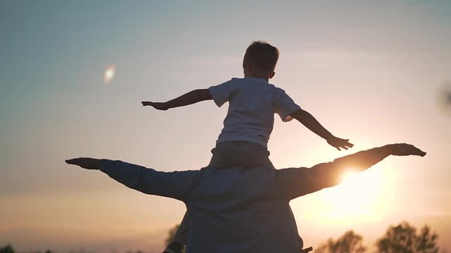Child sits high on shoulders of father. Boy stretches arms like wings at sunset. Outdoor family silhouette moment. Father supports child in sunset light. Kid balances arms in open air scene.