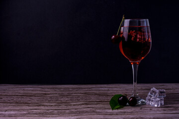 Glass of red cherry drink with ice cubes and fresh cherries on wooden table against dark background.
