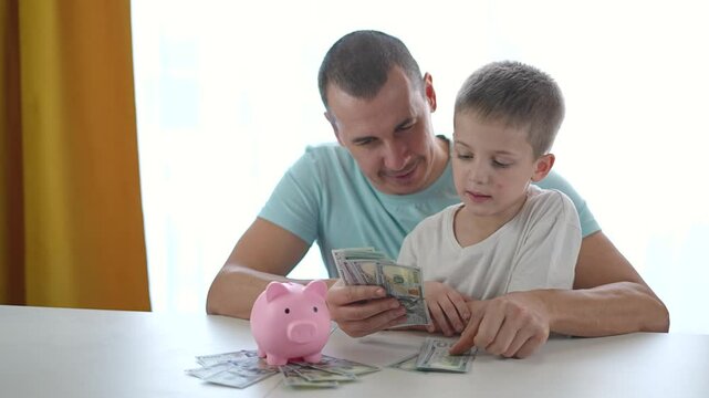 Father teaches boy to count dollar bills while sitting together at table. Father repeats saving instructions using money. Boy learns practical finance as father explains saving again and again