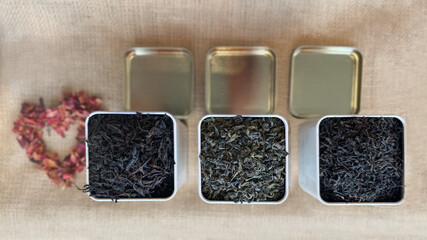 View from above of square metal jars filled with organic loose leaf tea, arranged on a burlap surface. A small decorative heart made of dried flowers adds a rustic and natural accent.