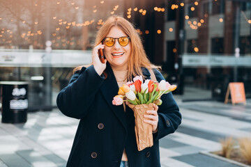 Woman smiling holding tulips talking on phone outdoors. Happy girl posing outdoors. Spring portrait...